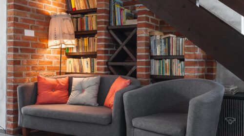 Office space featuring a brick wall under a staircase, the wall features book shelves with books and a lamp, a grey love seat with three throw pillows and a grey chair are sitting in front of the shelves